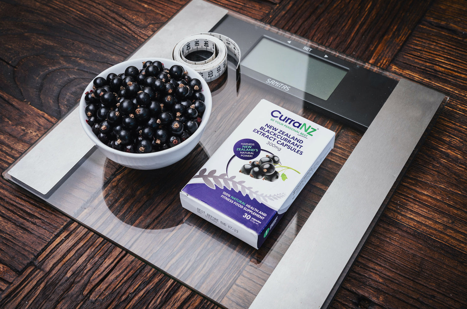 A packet of CurraNZ® supplements on a scale with NZ blackcurrants and a measuring tape.
